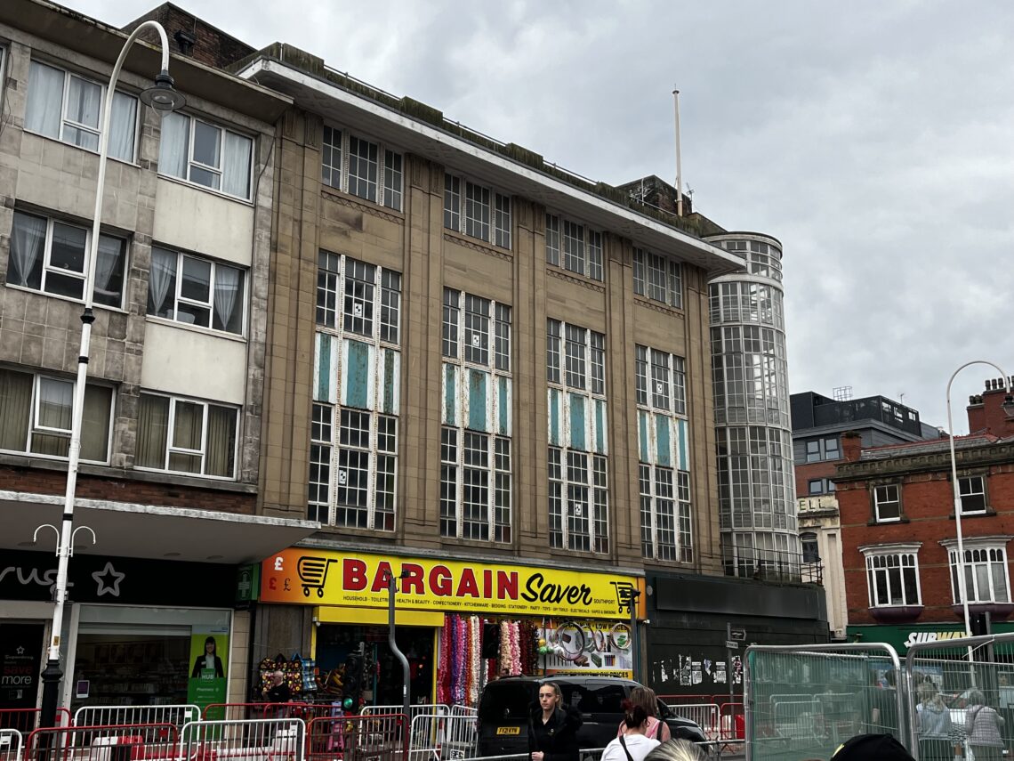A 4 storey department store building with some touches of art deco style. On the corner, there is a round window with a staircase visible.