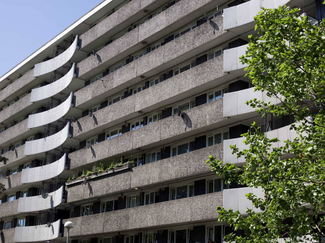 A shot of a 9 storey housing estate building shot at an angle of about 30 degrees to the face of the building. Each storey is fronted by a rough textured concrete walkway, and at equal intervals ribbed balconies made of an opaque white material, possibly also concrete, curve outwards. Residents' satellite dishes cast shadows in the spring sun. In the foreground, a leafy tree covers most of the first set of balconies.