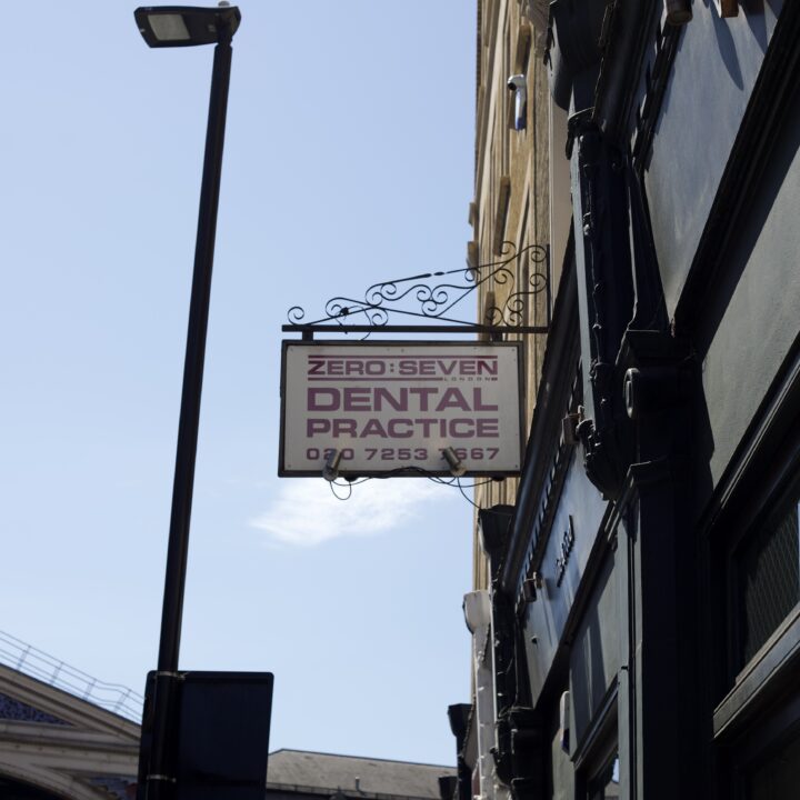 A sign reading Zero Seven London Dental Practice set in a wide, bold sans serif font. The words Zero and Seven are separated with a colon and feature a thick solid border above and below. They are on one line, with the word London in a much smaller font, inset into the border underneath. Underneath that, it says Dental Practice and the phone number, in a larger font across 3 lines.