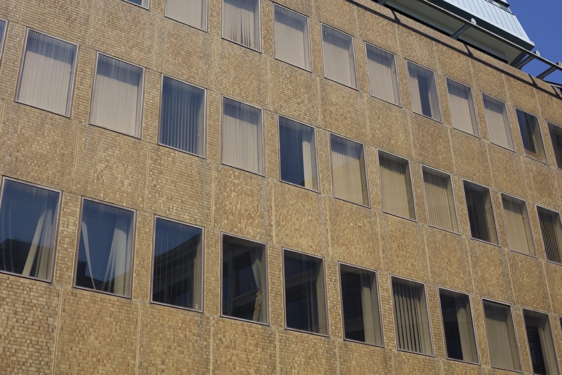 A close up of 3 storeys of windows, with a little bit of the sky visible in the top right corner. The building itself is clad with different shades of brown tile. The windows span the width of the building at equal intervals, and are darkened. Behind them are dishevelled vertical blinds.