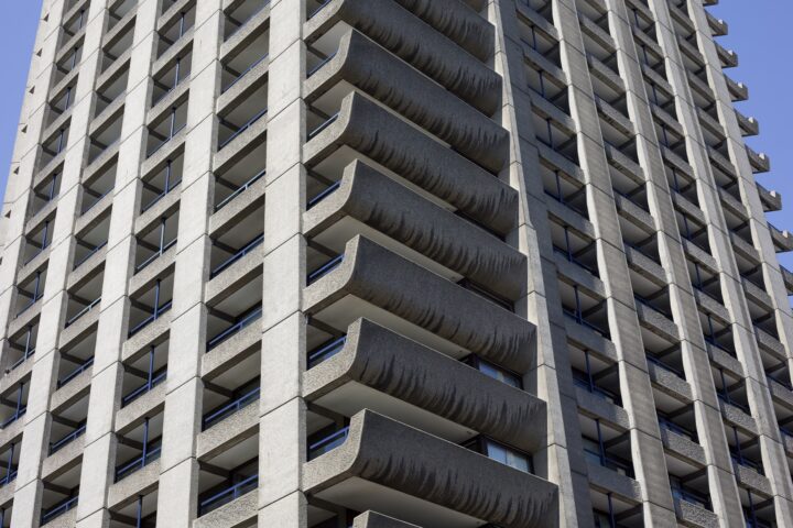 A sunlit ground-level shot of the corner of one of the towers at the Barbican Estate. In the centre of the image, the inflecting shape of the concrete balconies is captured. Several floors in the middle of the bulding are visible.
