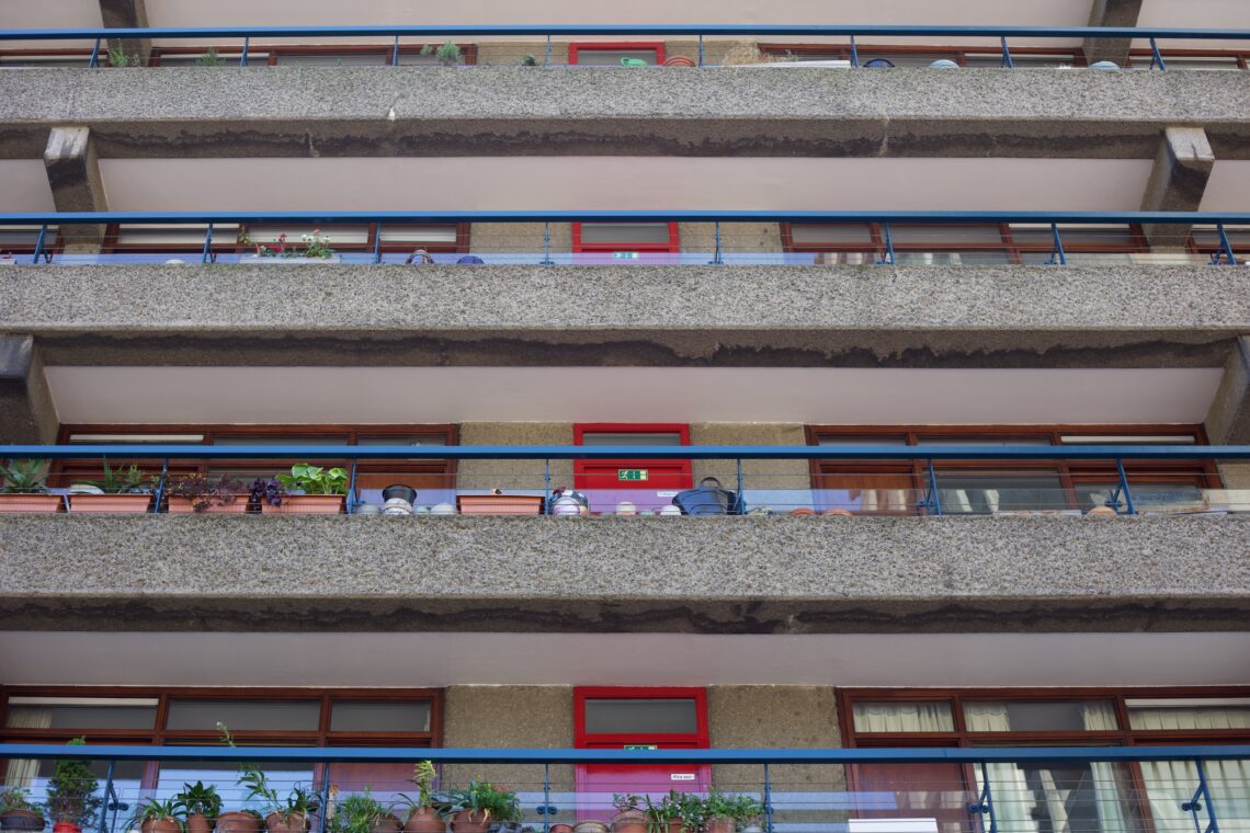 Close up of 4 concrete balconies, with blue metal and glass balustrades. Behind each, are some rectangular brown windows and bright red doors. The shot is taken from ground level, looking up at the balconies, filling the image geometrically.