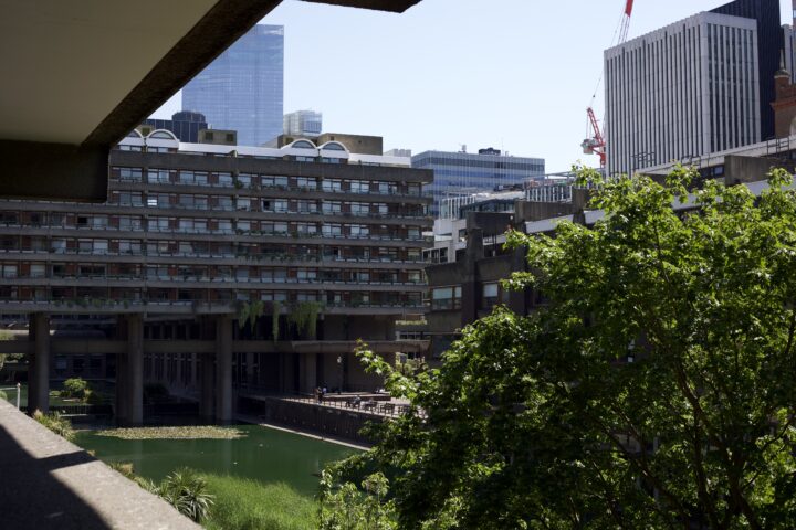 A shot from one of the balconies at the Barbican estate. In the very front of the shot, the concrete balcony is out of focus. Further out, there is a tree, a pool green with algae, and even further back, one of the apartment buildings and the modern glass buldings of the City of London in the background.