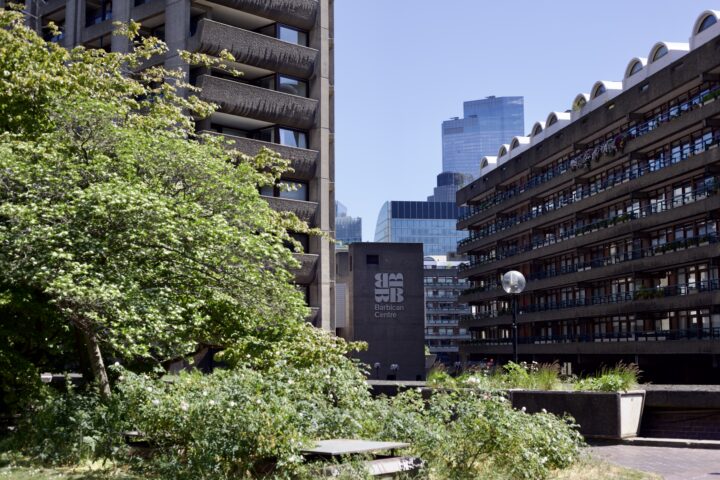 A shot of the Barbican estate, featuring greenery in the very foreground, followed by Barbican buildings further back, and the glass buildings of the City of London in the very background.