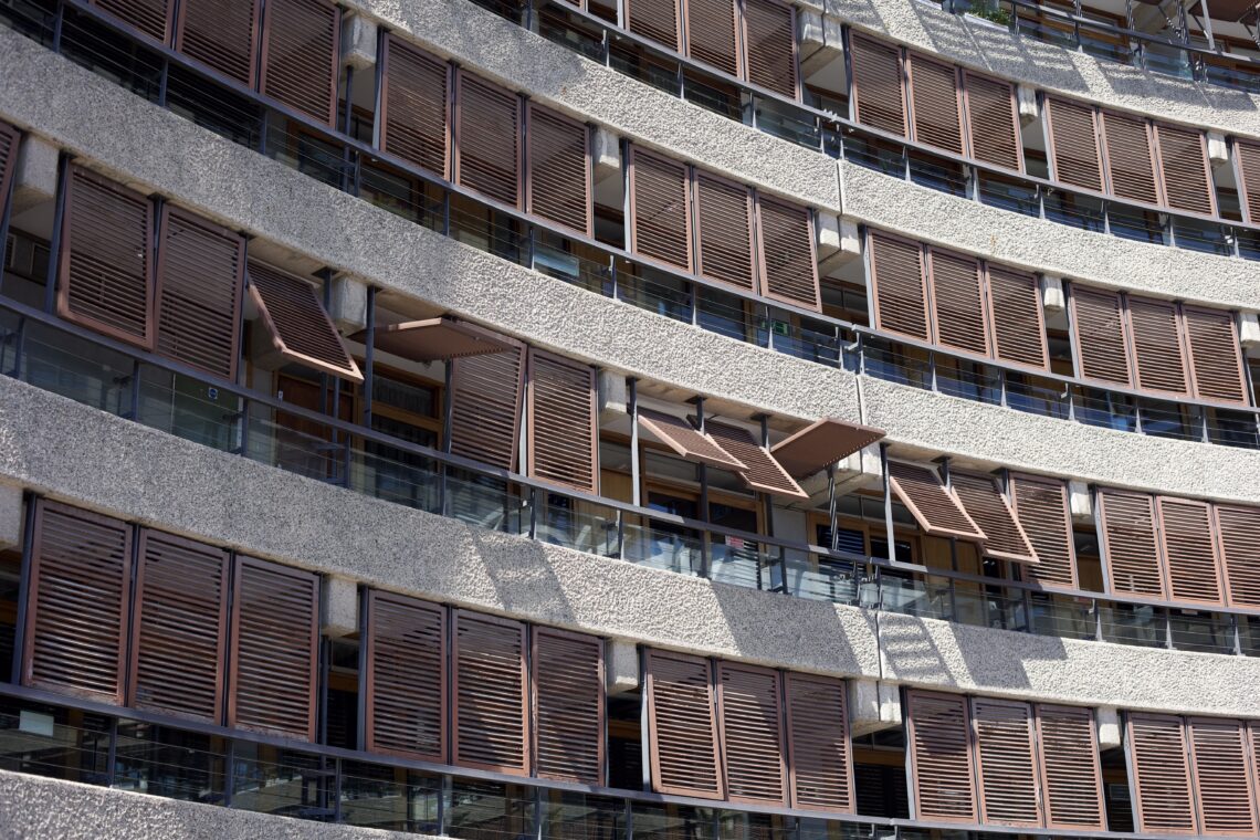 A sunlit shot of balconies covered with shutters on a curved building at the Barbican Estate. Most of the shutters are closed, but on the floor which is in the middle of the shot, several of them are open to differing degrees.