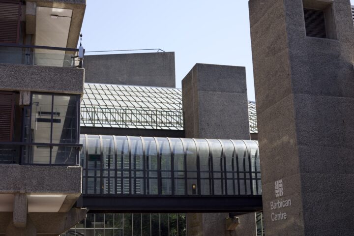A shot of the Barbian Estate, with multiple parts of it visible. In the foreground, on the left, the edge of one of the buildings with corridors and stairways visible, as well as a concrete ventilation shaft on the right. Across the middle of the shot, a covered glass walkway. In the back, another concrete shaft and the conservatory.