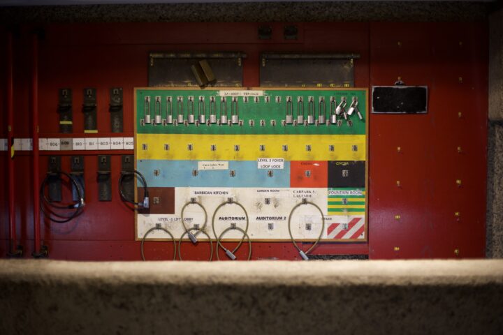A multi-coloured contraption for holding bike locks, mounted against a red wall. It has several sections, all of different colours, with labels in different fonts and of different ages. Some have bike locks attached to them.