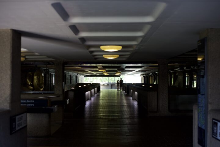 A shot of one of the covered walkways of the Barbican. It is fairly dark, with light only coming in at the back of the image, where silhouettes of two people can be seen standing.