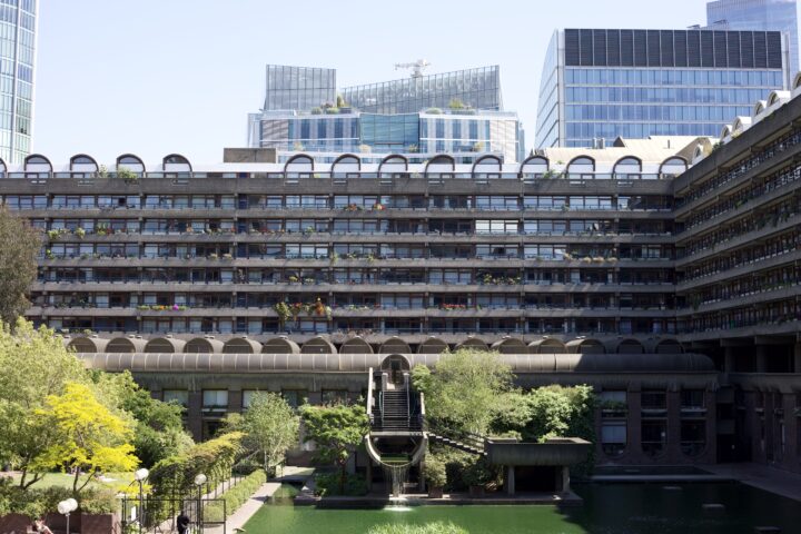 A wide geometric shot of flats at the Barbican estate. In the background, more modern glass buildings of central London. In the foreground, a fountain with water flowing into the pool beneath.
