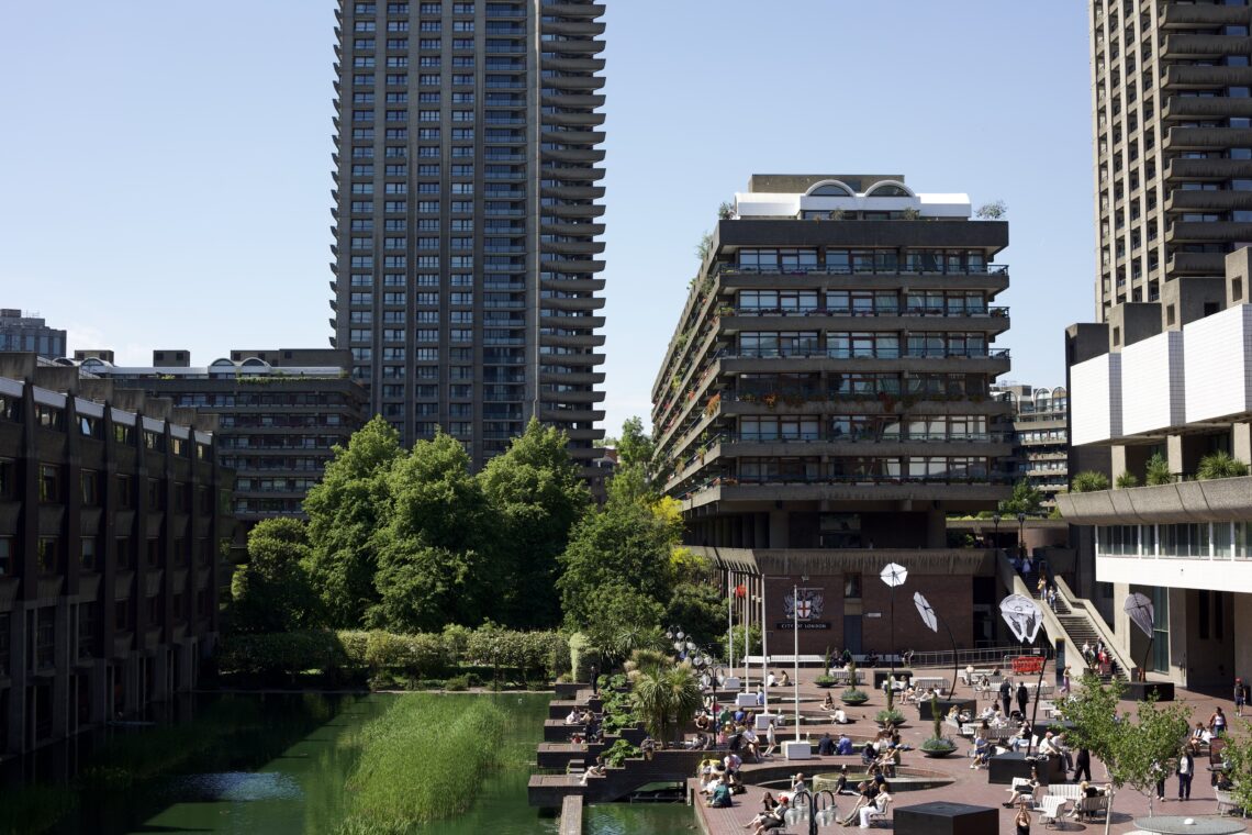 Wide shot showing many features of the Barbican estate. From left to right: Guildhall School of Music and Drama, an algae-green pool, a tall tower, a shorter tower with view of its balconies, and the main Barbican library building.