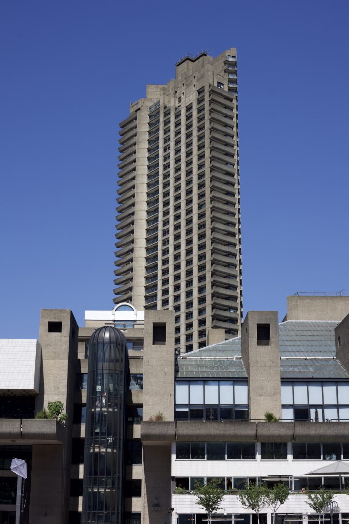 In the foreground of the picture, the Barbican Centre is visible, with a white-clad cafe to the right, a round staircase clad in darkened glass, and 3 concrete ventilation towers. In the background, against a dark blue sky, a concrete tower block rises.