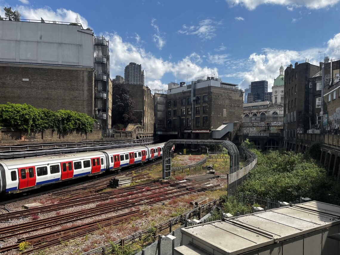 A view of the backs of various London buildings, with a Tube line with a modern Tube train running on it. At the back, centre-left, two of the Barbican towers are visible.
