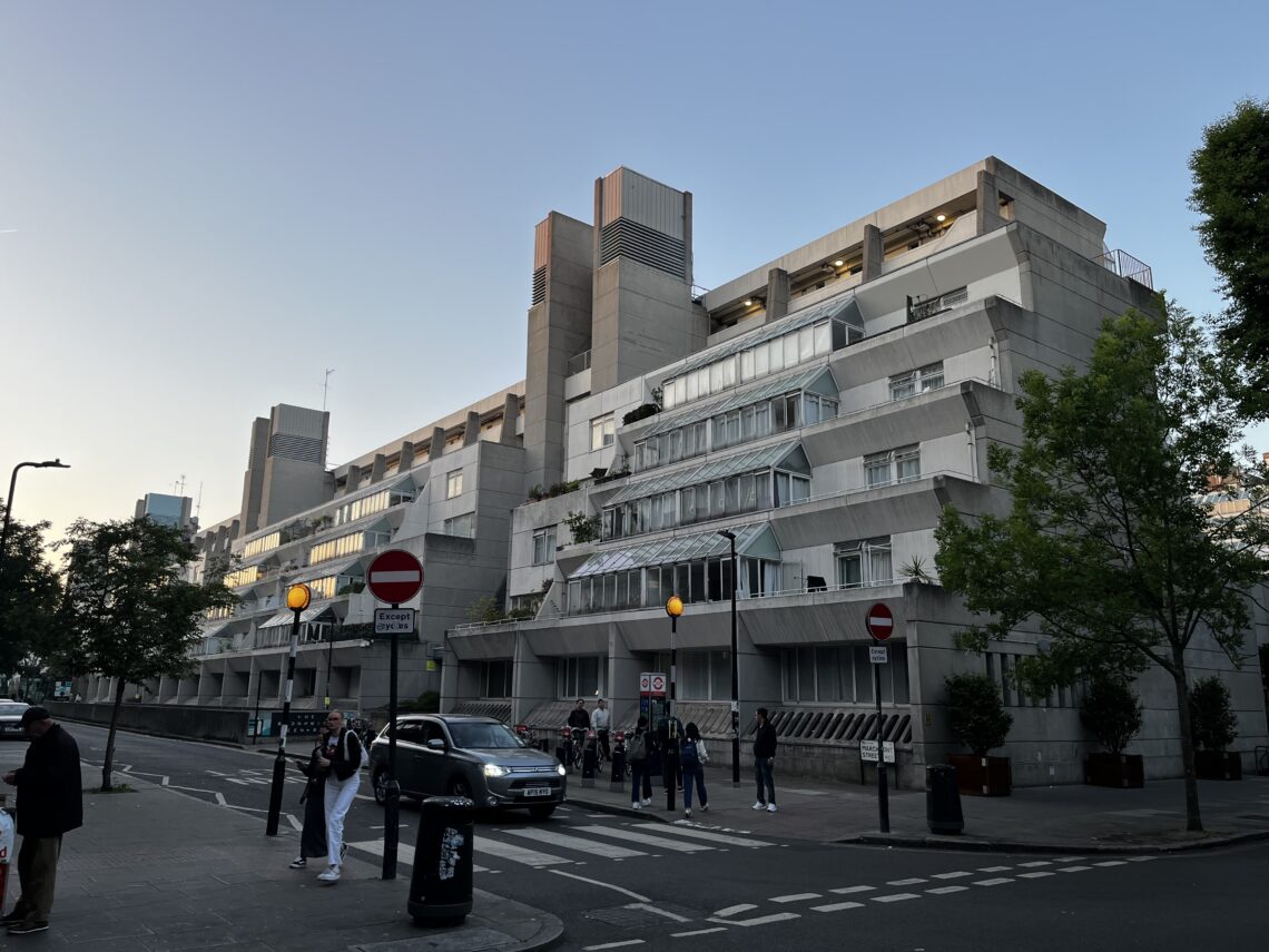 A photo of the Brunswick Centre taken at a 45 degree angle. It is a concrete ziggurat-style structure, painted white, with small conservatory-like glass balconies on each floor. The sun is setting, and people are crossing the zebra crossing on front of the building.