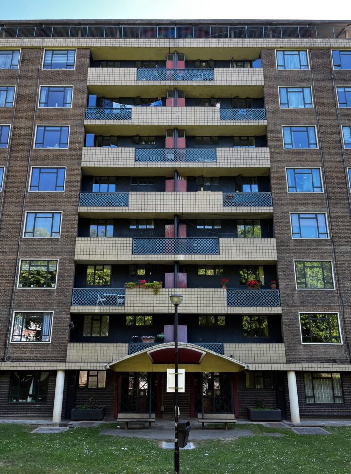 Ground level shot looking up at an 8 storey building, focusing on the balconies. These alternate with tiles at the sides and latticed fencing in the middle, and vice versa. The balconies are inset into the building and either side of them is a grid of clean, PVC windows.