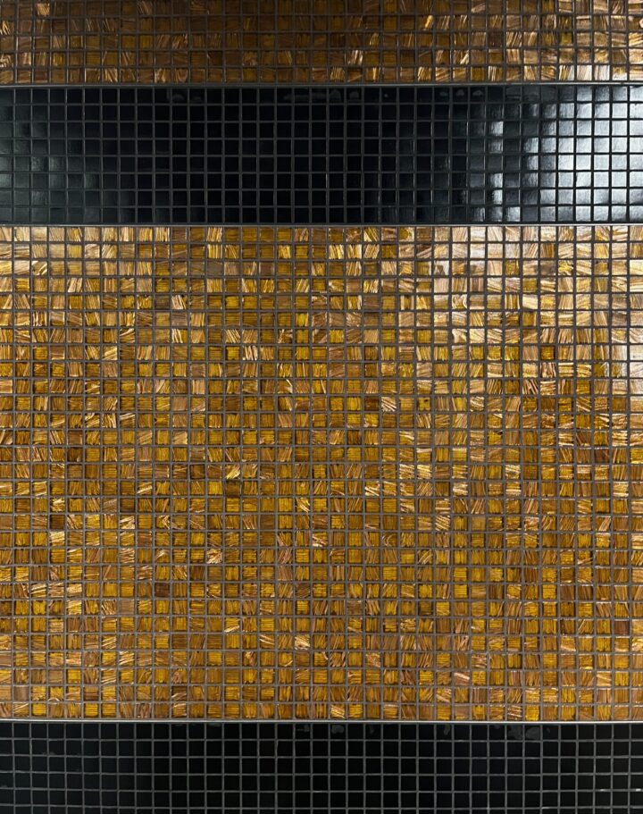A close up shot of small square tiles. They are primarily amber, and as they reflect in the light you can see the pattern embedded within them goes in different directions. There are also two bands of black tiles, one towards the top, and one at the very bottom.