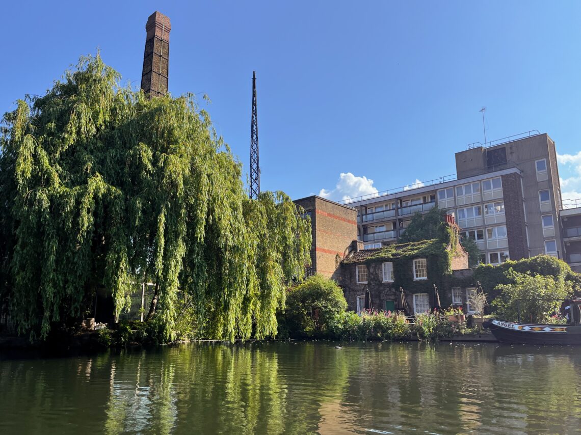 Sunny day on the Regent's Canal, taken from on the water. The foreground features a large, lush green tree. In the middle, there are some 19th century buildings, including a mill and cottage. In the background there is a 1960's mid-rise housing block, all set against a clear blue sky.
