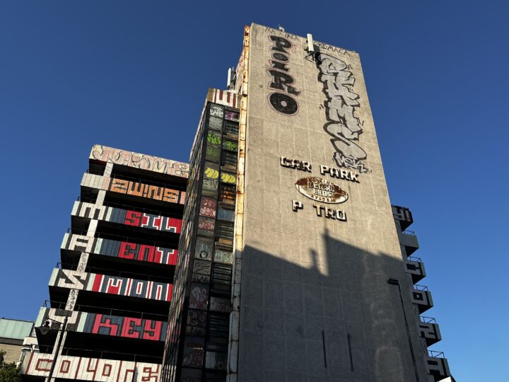Side of multistorey carpark, taken from ground level. The side of the concrete is made out of concrete panels, and the top part of it is graffitied. Below that, a rusty sign which reads: CAR PARK P_TRO_ - Meyers Bros Parking System. The rest of the image features the graffitied tiers and stairway of the car park, with the dark blue late afternoon sky in the background.