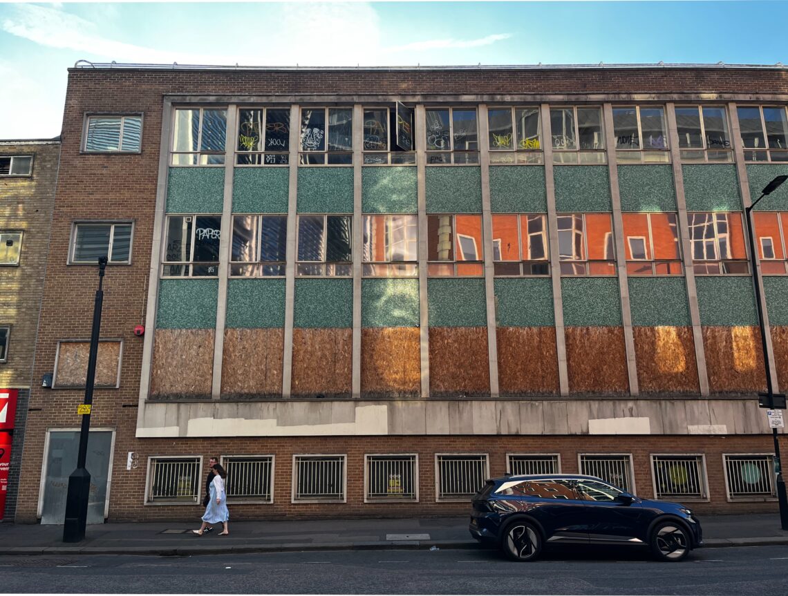 Brick building with a concrete brick and window facade. The first floor features grates, as if for a semi-underground car park. All windows on the floor above are boarded up. Windows on the higher floors are not boarded up, but are largely graffitied.