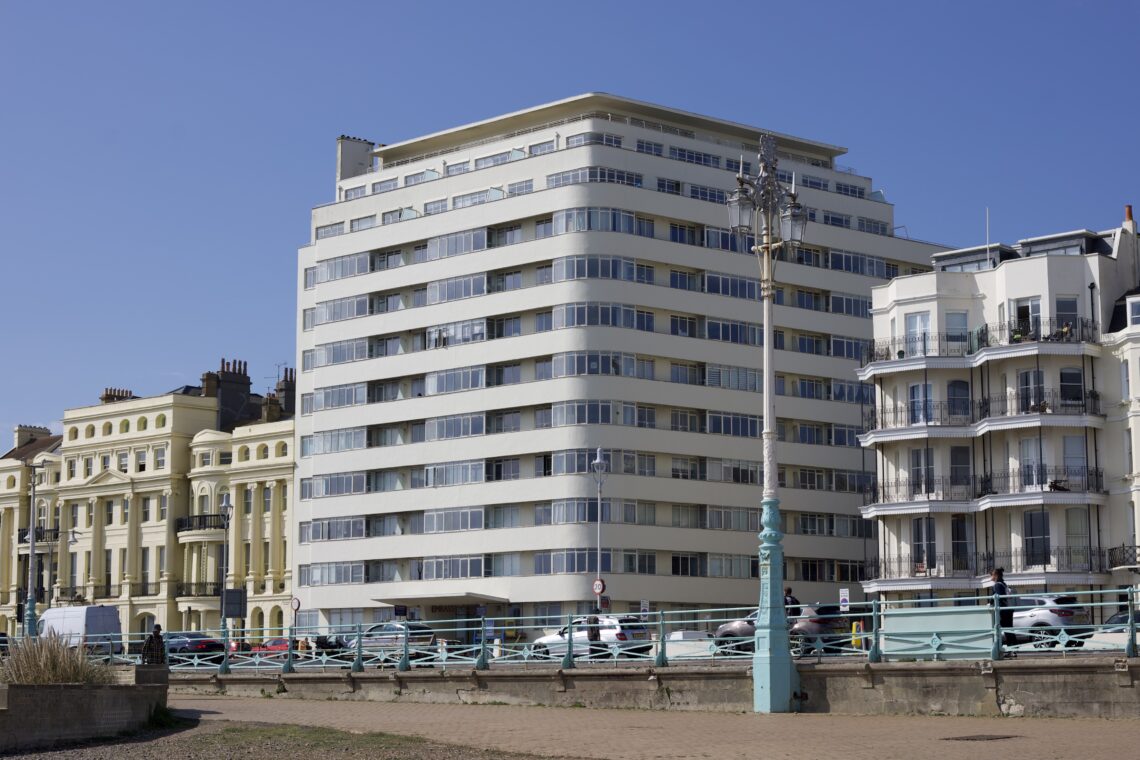 A corner-on shot of an art deco building with a square mass, painted white. It is surrounded by smaller, younger buildings. The main body of the building has 9 storeys, with the top 3 storeys receding back from the main body of the building. Each floor of the building features windows, with interspaced balconies.