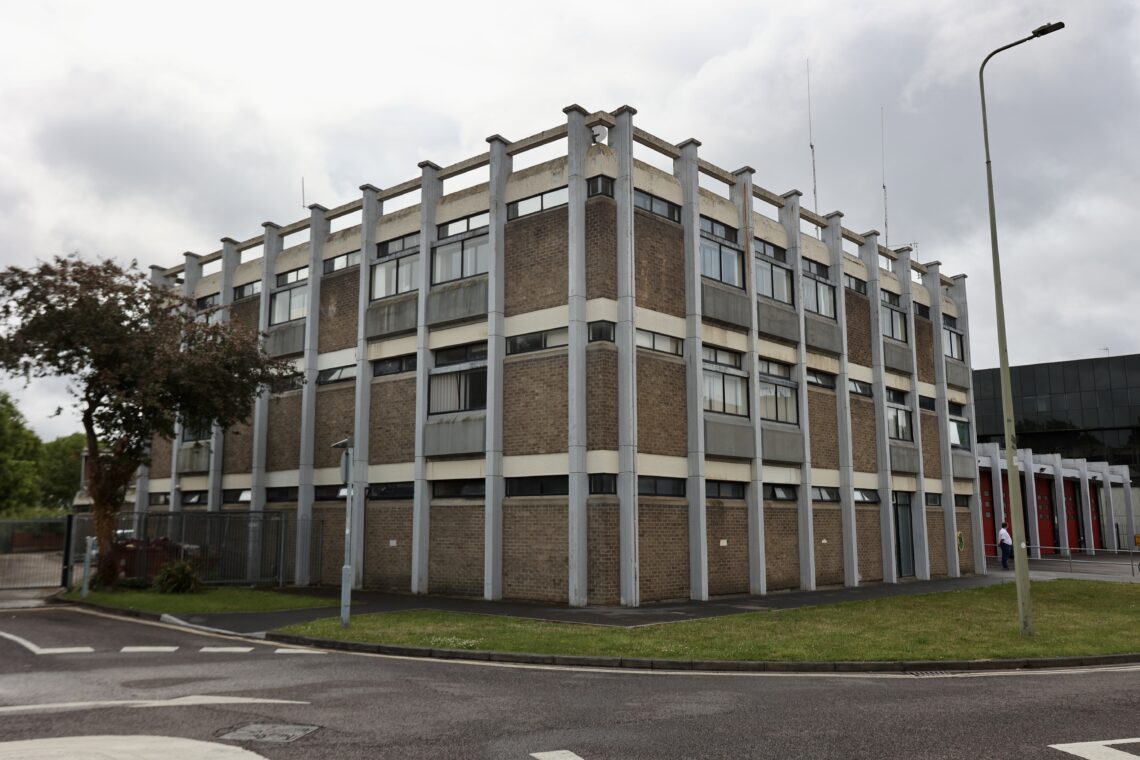 Corner-on shot of a 3 storey concrete framed brick building. In the background of the shot, the building extends into what is clearly recognisable as the red shutters of a fire station, separated by an external concrete frame. The bulding is set against a cloudy grey sky, with some grass and a road in the foreground.