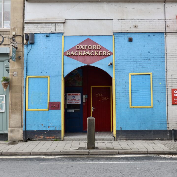 The photo is set on a city street, capturing the street, pavement, and a single bollard in the middle of the shot, and shows the entrance to a hostel. A square section of a large, white, unassuming brick wall is painted bright baby blue, with yellow and dark red detailing. A dark red diamond above the make-shift entrance arch reads OXFORD BACKPACKERS in a whimsical font.
