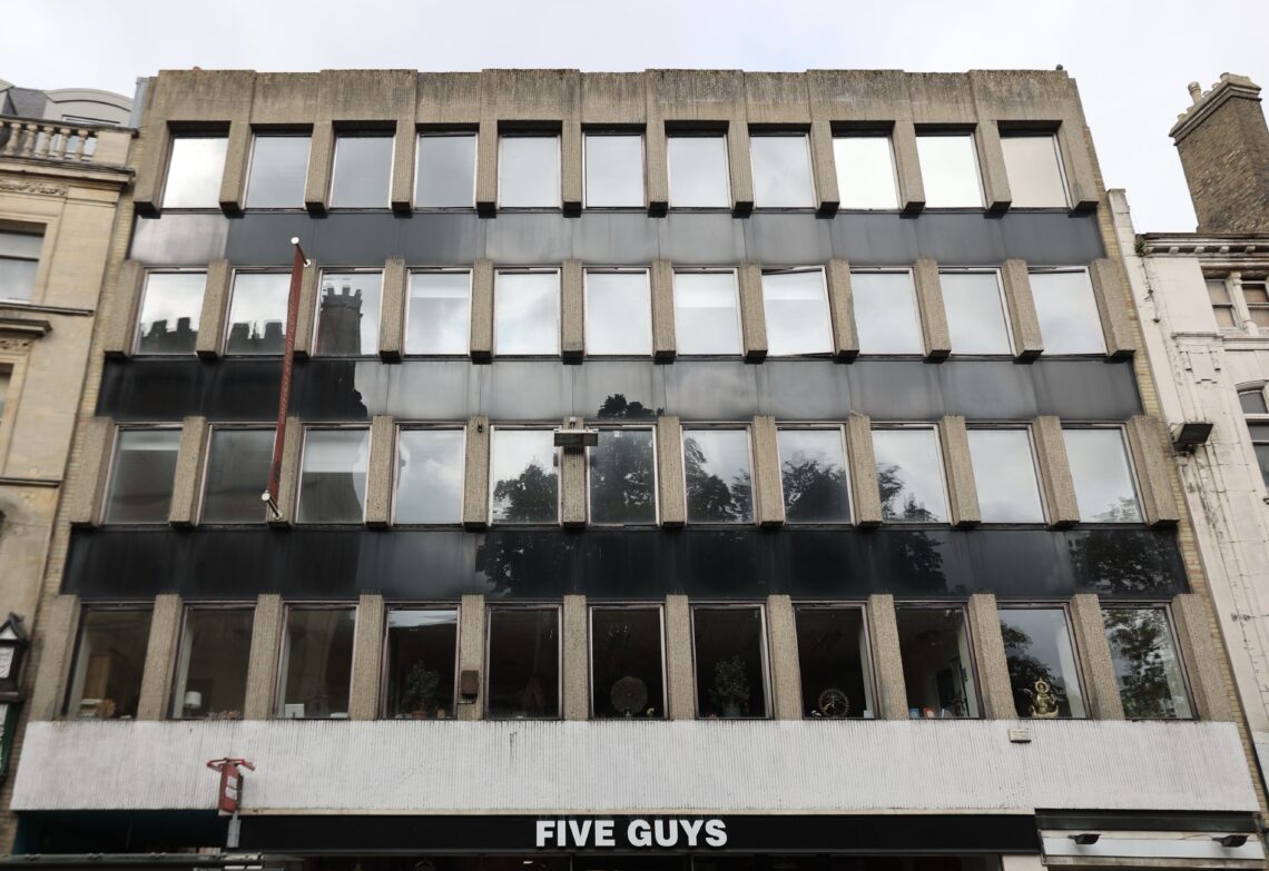 A beige, modernist building with five rows of square windows is shown. The ground floor has a FIVE GUYS sign. Reflections of trees and a church tower are visible in the windows. The sky is cloudy.
