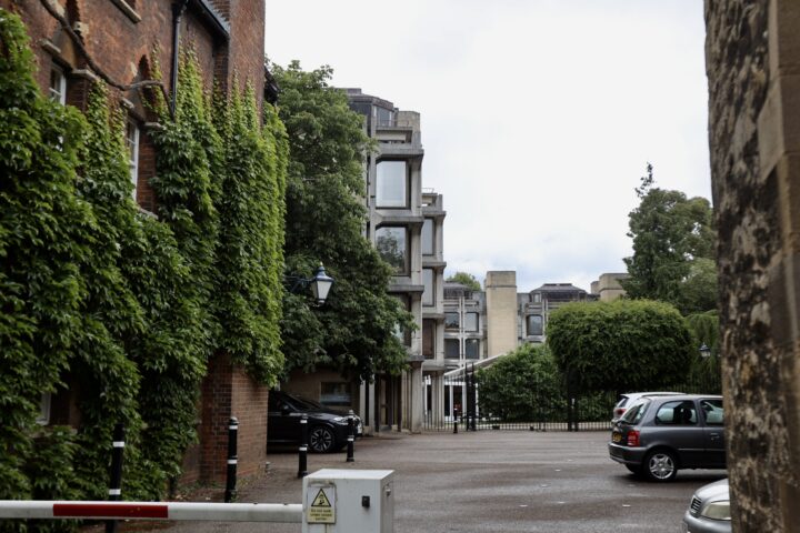 A small car park with several cars is surrounded by older brick buildings with ivy on the left and modern windowed buildings ahead. Trees and greenery line the area. A white barrier and a “danger” sign are visible in the foreground. The sky is overcast.