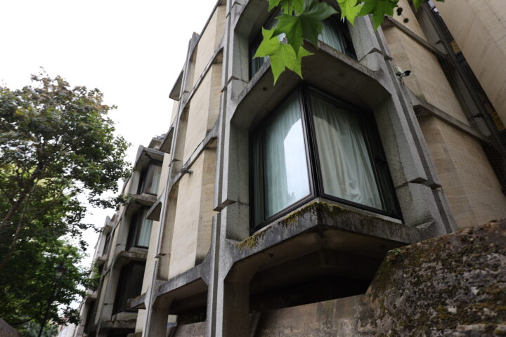 A close-up of a brutalist building features large concrete bay windows with black frames and curtains drawn inside. Moss grows on ledges, and leafy green trees are visible on the left side, adding contrast to the angular concrete structure.