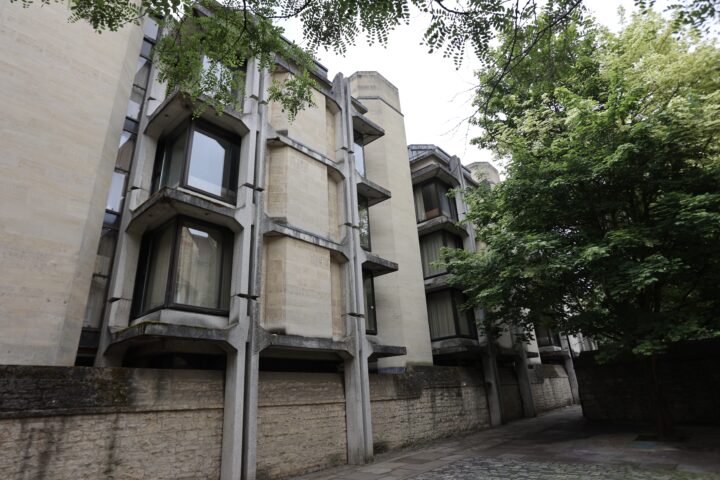 A modern concrete and stone building with angular windows and vertical supports stands beside a cobblestone path, bordered by a stone wall. Lush green trees partially obscure the structure, and the sky is overcast.