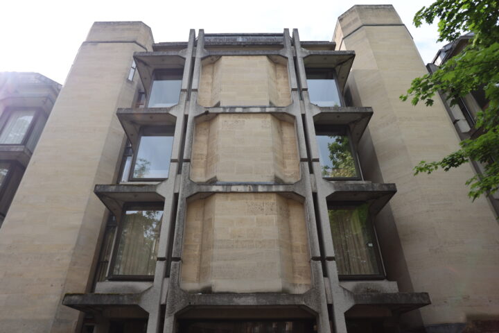 A concrete and stone building with tall, narrow windows and vertical, ribbed concrete supports on the facade. The structure has a modern, brutalist style and is partially shaded by a leafy tree on the right. Sunlight appears from the upper left corner.