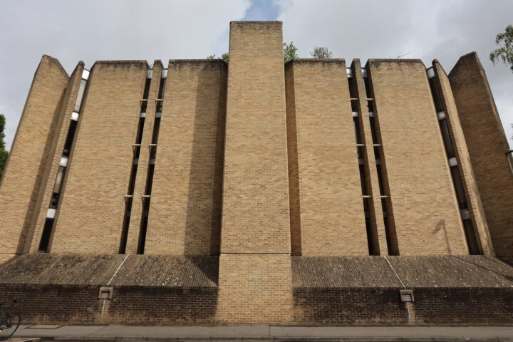 A tall, fortified-looking building made of yellow-brown brick, with multiple vertical columns and narrow, slit-like windows. The geometry is angular and brutalist. The sky above is cloudy, and a bicycle is parked at the far left.