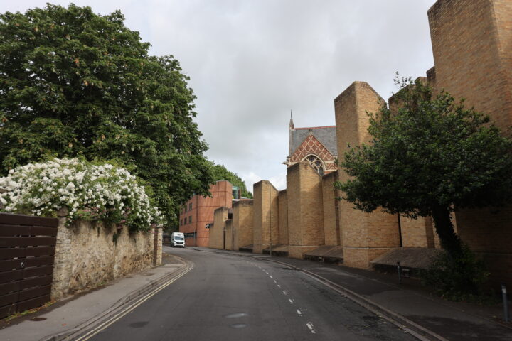 A narrow, empty street curves alongside modern brick buildings with distinctive vertical buttresses. On the left, a stone wall with blooming white flowers and leafy trees contrasts with the architecture. The sky is mostly cloudy, and a white van is visible in the distance.