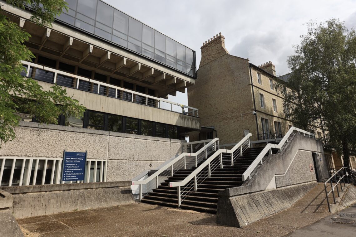 A modern concrete building with large windows sits next to an older brick building. A broad staircase with white railings leads up to the entrance. Several trees and a blue informational sign are visible, and the sky is partly cloudy.