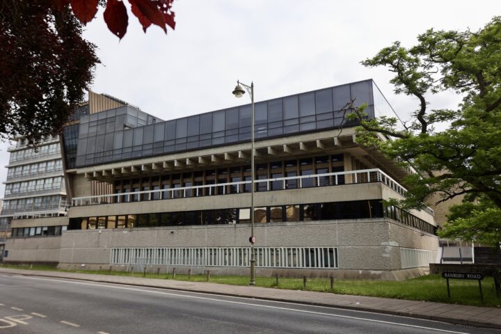 A modern, multi-storey building with large glass windows and concrete walls stands beside a road. A street lamp is in front, green trees frame the right and top left, and a sign reads Banbury Road at the corner. The sky is overcast.