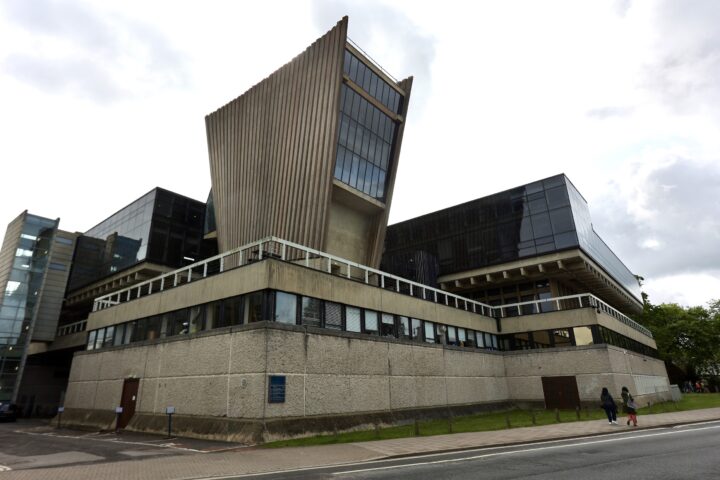 A large, modern building with angular, jutting concrete and glass structures. The lower level is textured concrete, while upper floors feature large dark glass windows. Two people walk on a pavement beside the building on a cloudy day.