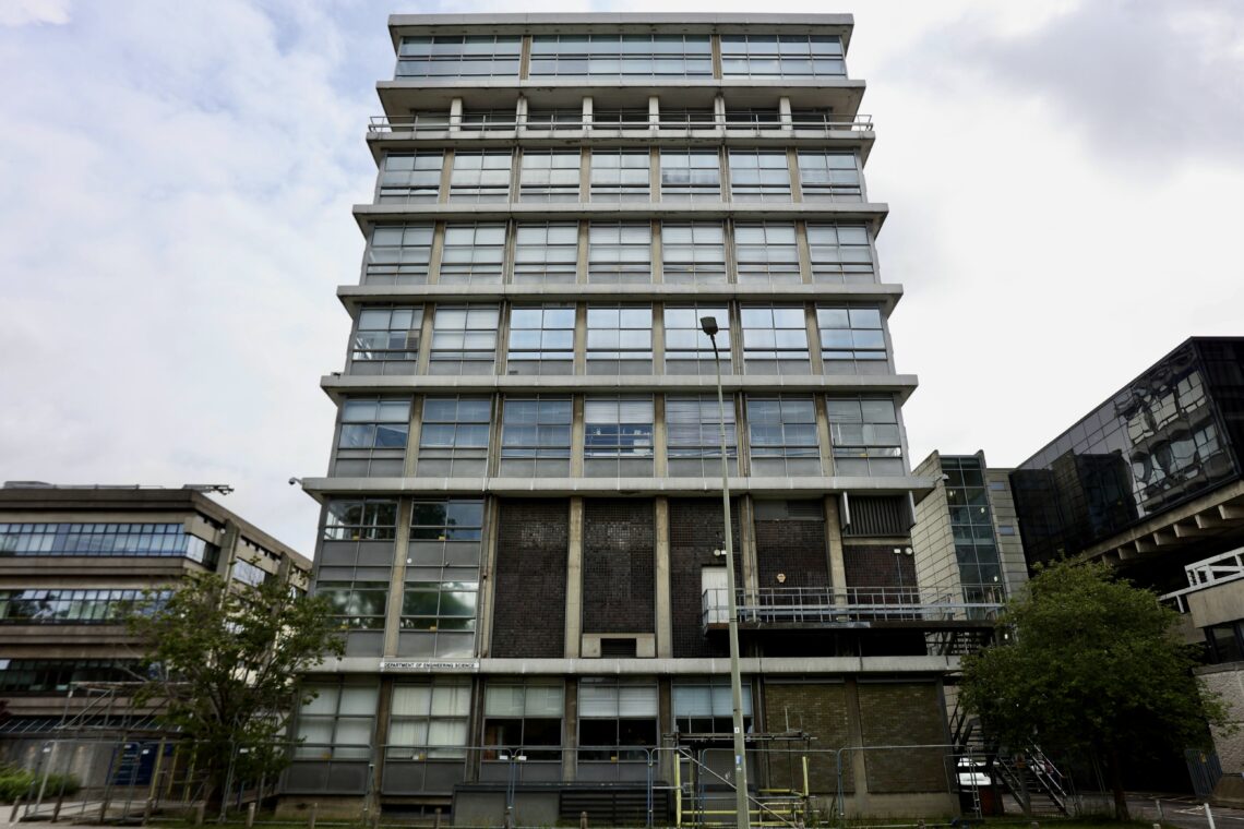 A large, square, concrete and glass building with multiple floors, seen from the front. It has many windows, a flat roof, and a central pole with lights in front. Trees and other buildings are visible on either side under a cloudy sky.