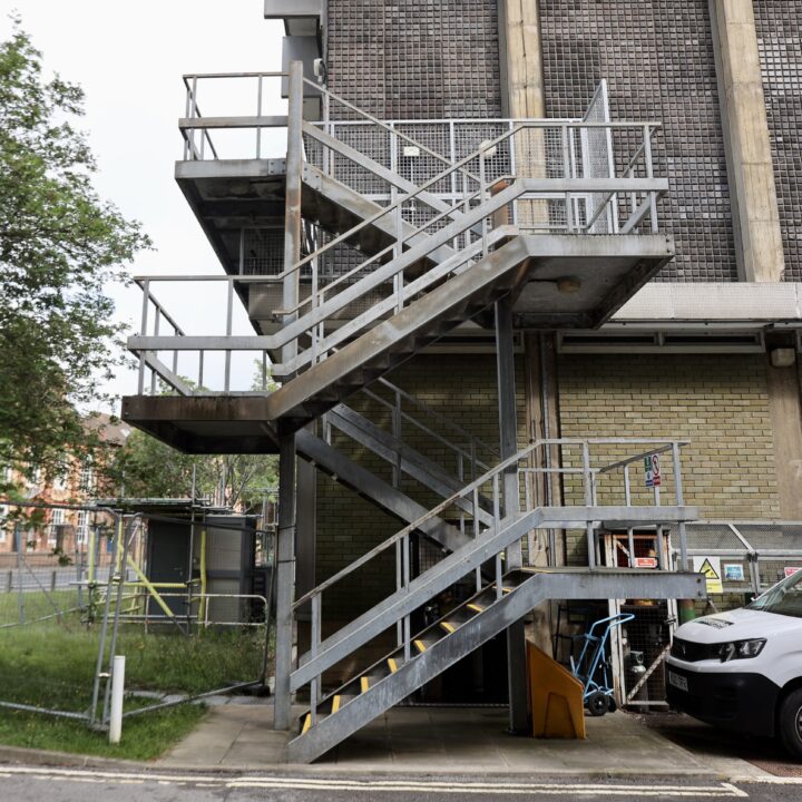 Metal exterior staircase with three flights and railings attached to a brick building. Yellow caution lines on the middle stairs. A blue bicycle and various equipment are underneath. Grass, trees, and a fence are on the left; pavement and a road are in front.
