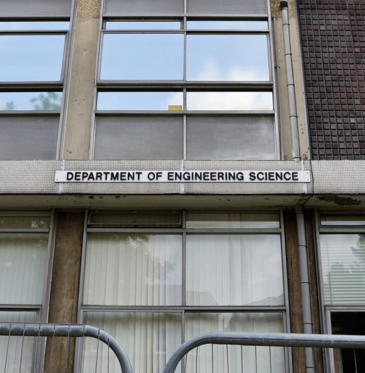 A rectangular sign reading “Department of Engineering Science” is mounted above large windows on a concrete and brick building. Metal barriers are visible in front of the entrance, and reflections can be seen in the upper windows.