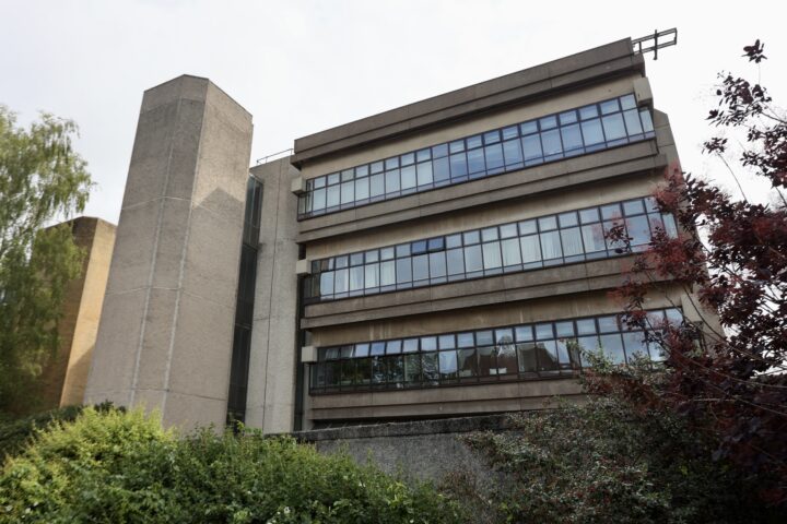 A large concrete building with four storeys of horizontal windows, each floor lined with continuous glass panels. The structure has a tall, plain stairwell tower on the left and is surrounded by dense green foliage and some red-leaved bushes in front.
