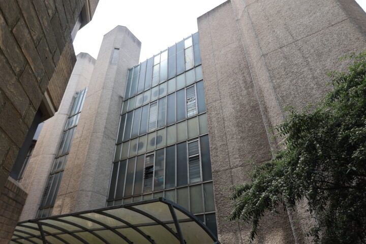 A tall, modern concrete building with narrow vertical windows and textured walls. The facade features glass panels with horizontal metal dividers. A transparent, curved canopy is in the foreground, and leafy branches appear on the right side.