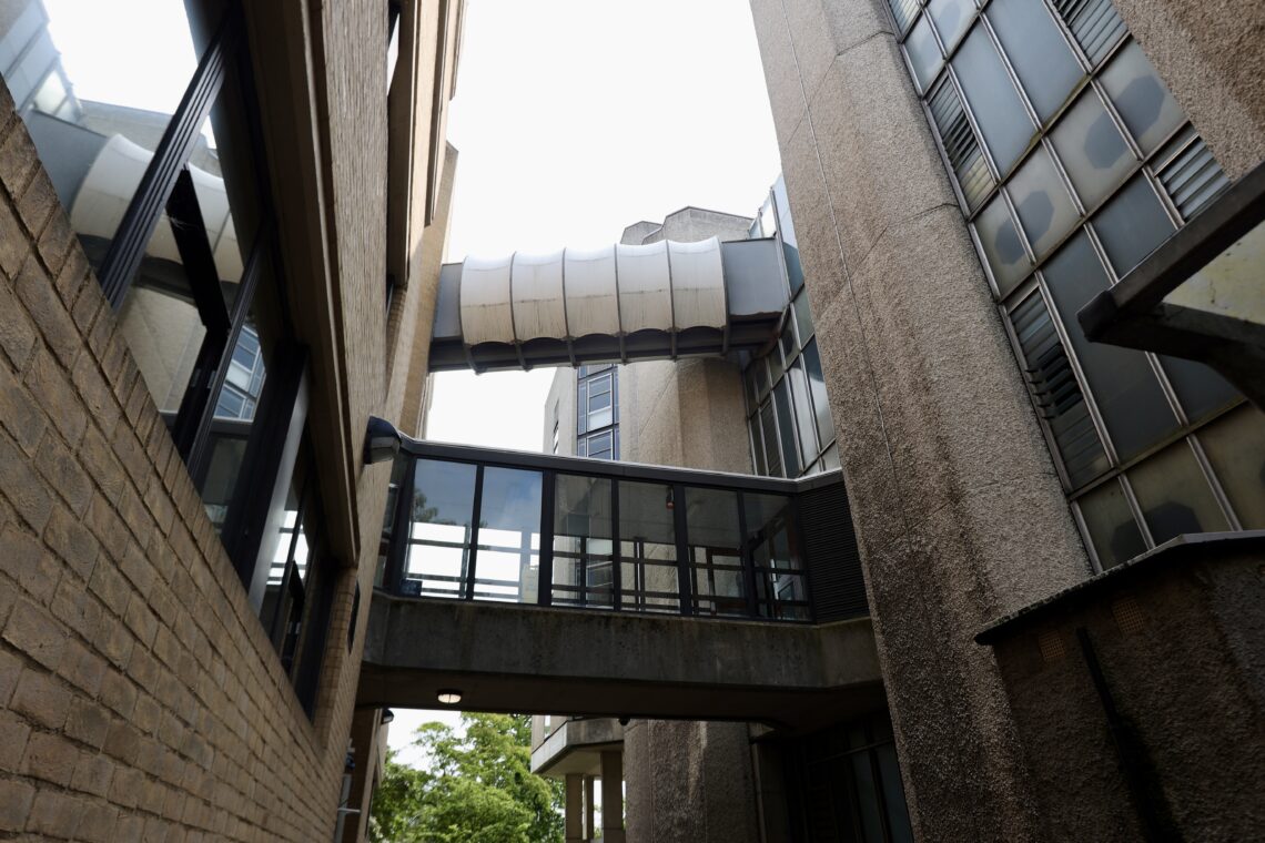 View looking up between two Brutalist buildings connected by two elevated walkways, one with glass panels and another with a curved metal covering. The buildings have rough concrete and brick textures, with some windows and vents visible. Trees can be seen below.