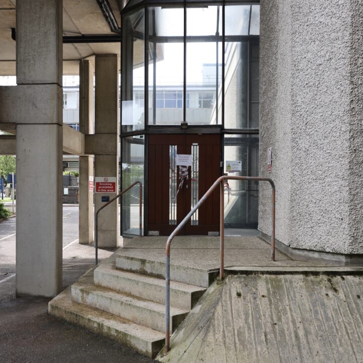 Concrete steps with a metal railing lead to a glass entrance door framed in dark red, set in a modern concrete building. The wall on the right is textured. Large windows above reflect another building. A ramp runs beside the steps.