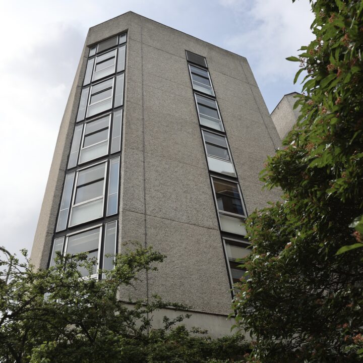 A low-angle view of a tall, grey concrete building with narrow, rectangular windows. Green leafy trees partially frame the lower right and left edges, and the sky is partly cloudy in the background.