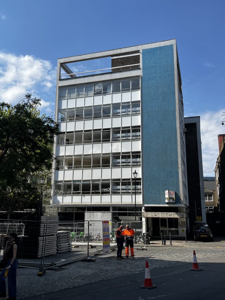 6 storey 1960's office building with an exposed concrete frame and a column of mosaic detailing above the main entrance, as well as a to let sign. The front face of the building features a geometric graph of thin-frame aluminium windows. In the foreground of the picture, roadworks.