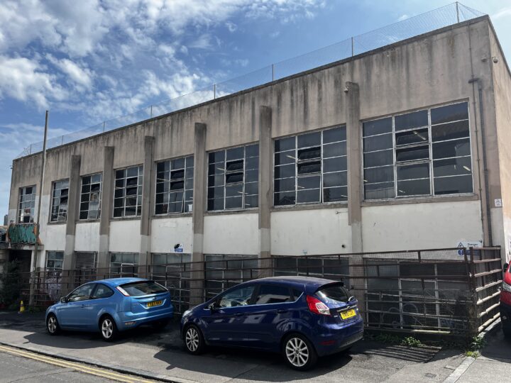 A plain concrete building featuring 7 sets of thin-framed windows with a concrete column inbetween each, except the 8th, which is a little further away with no column (and a passageway underneath). The lower part of the building has been painted white, although this is quite faded and irregular.