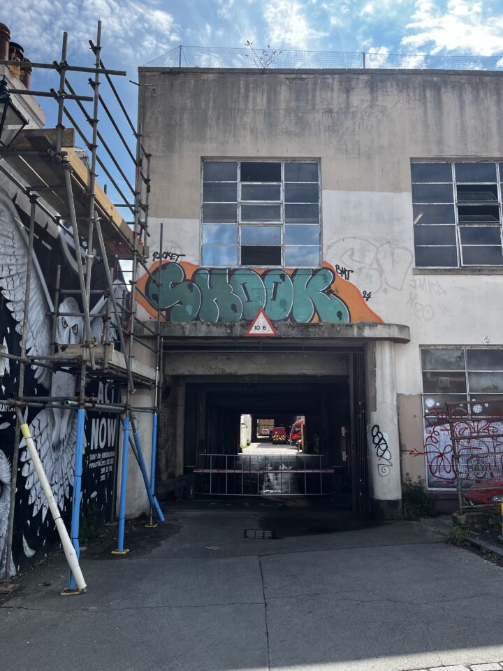 A front-on shot of a concrete building. The lower half of the building features a passageway, with a somewhat art deco canopy and column. Just above it is some graffiti, and a thin-framed window panelled window.