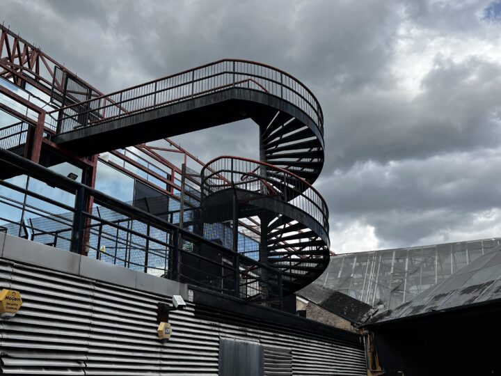 A spiral staircase at the side of a cascading multi-storey reflective glass pyramid. The staircase contrasts sharply against the cloudy sky, spiralling twice before a long narrow section leading into the building.