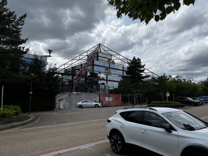 A more distant shot, set in a car park, of the cascading multi-storey reflective glass pyramid building set against a dramatic cloudy sky.