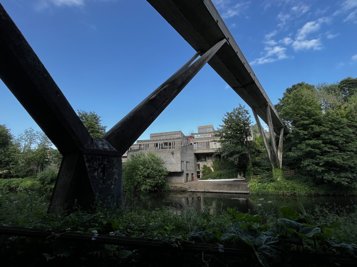 A large, angular concrete pedestrian bridge crosses over a lush river, supported by V-shaped pillars. Behind it, a modernist grey building with angular rooftops sits among dense green trees under a bright blue sky with scattered clouds.