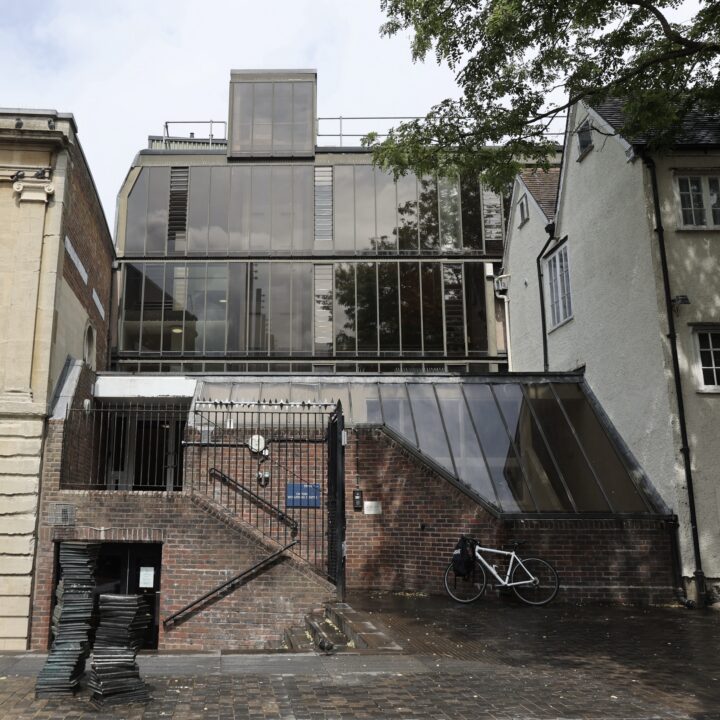 A modern building with large reflective glass windows and angled brick walls stands between two older structures. A staircase leads to a gated entrance, and a white bicycle is parked near the base. Tree branches hang over the scene on the right.